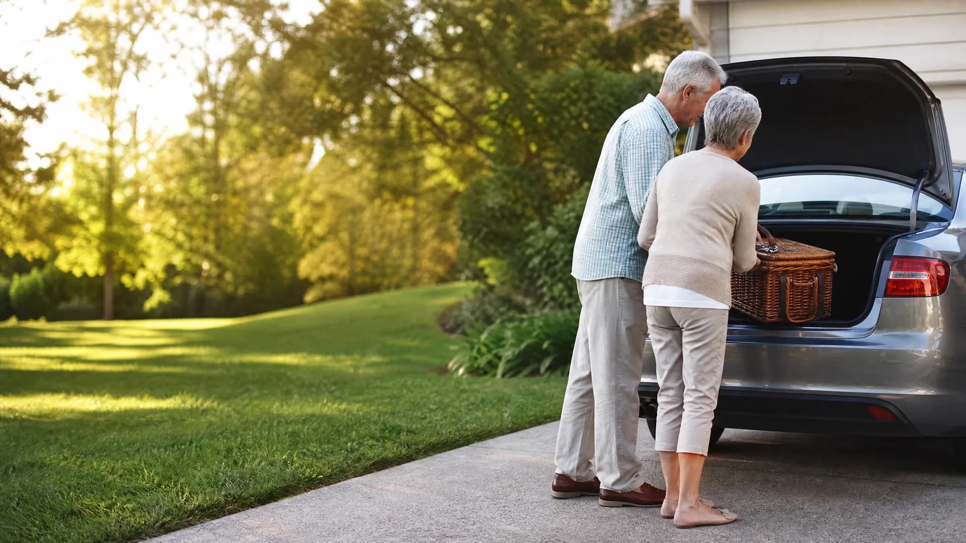 An older man and woman stand at the open trunk of a gray car in a driveway, loading a wicker basket, with a lawn and sunlit trees behind them.