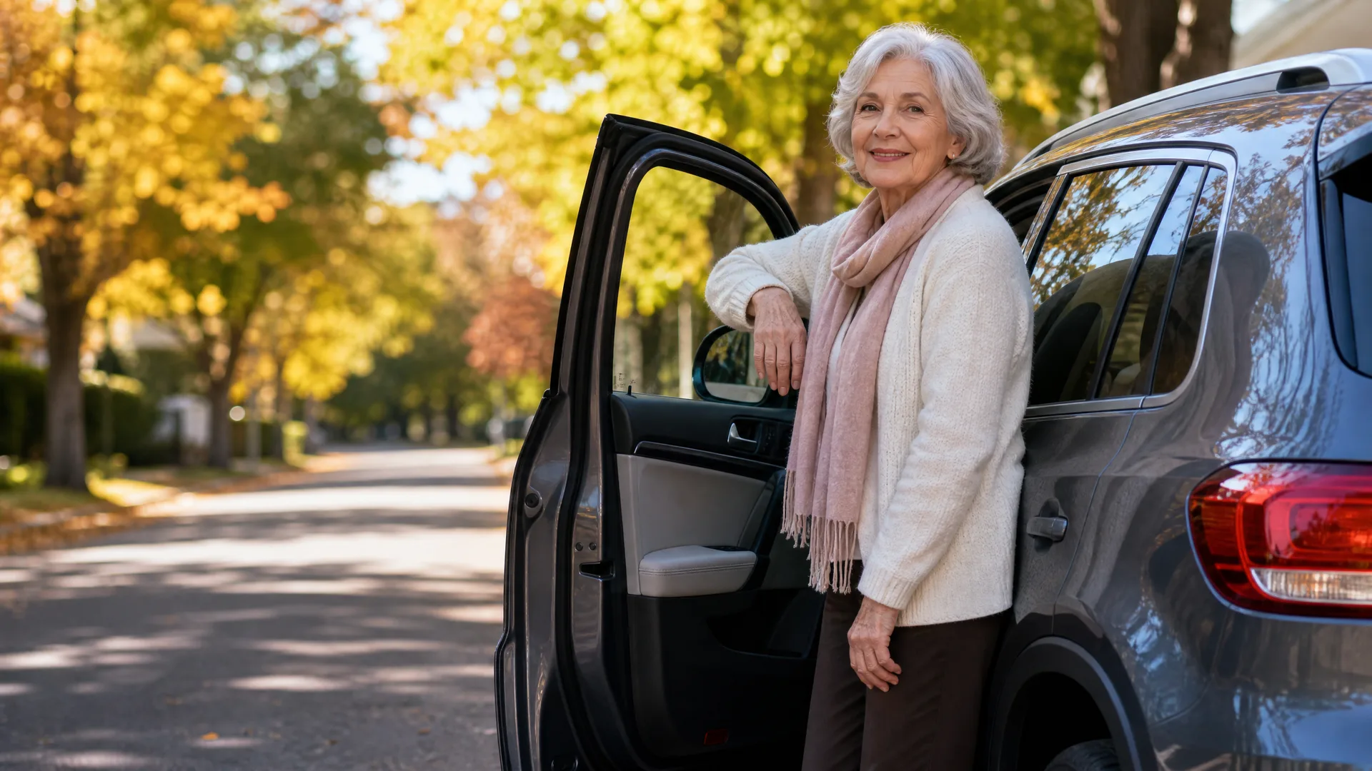 An older woman with white hair, wearing a cream cardigan and pink scarf, stands beside the open passenger door of a dark SUV on a tree-lined street with yellow autumn foliage.
