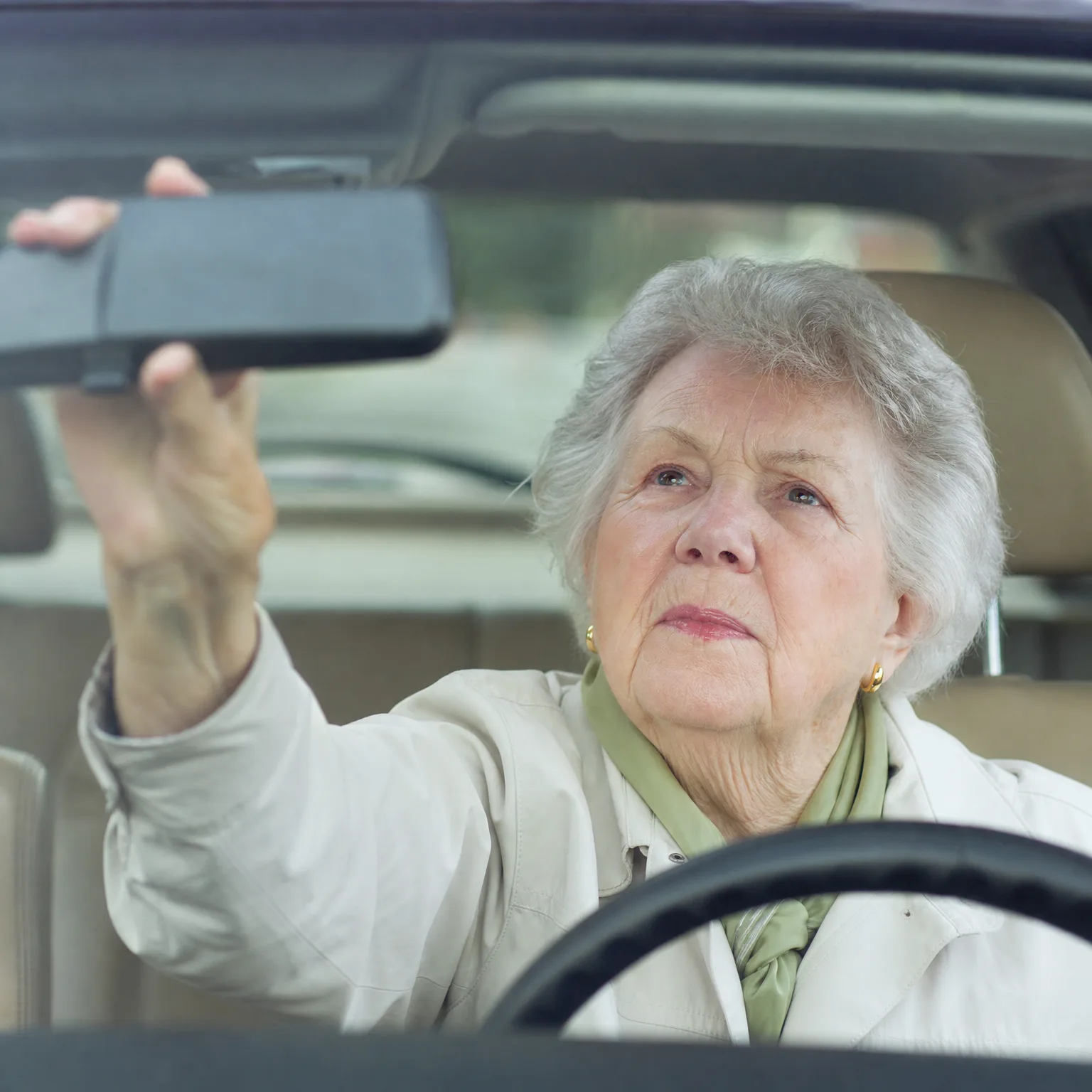 An older woman with short gray hair, wearing a light jacket and green scarf, sits in a car's driver seat and reaches up to adjust the rearview mirror, viewed through the windshield.