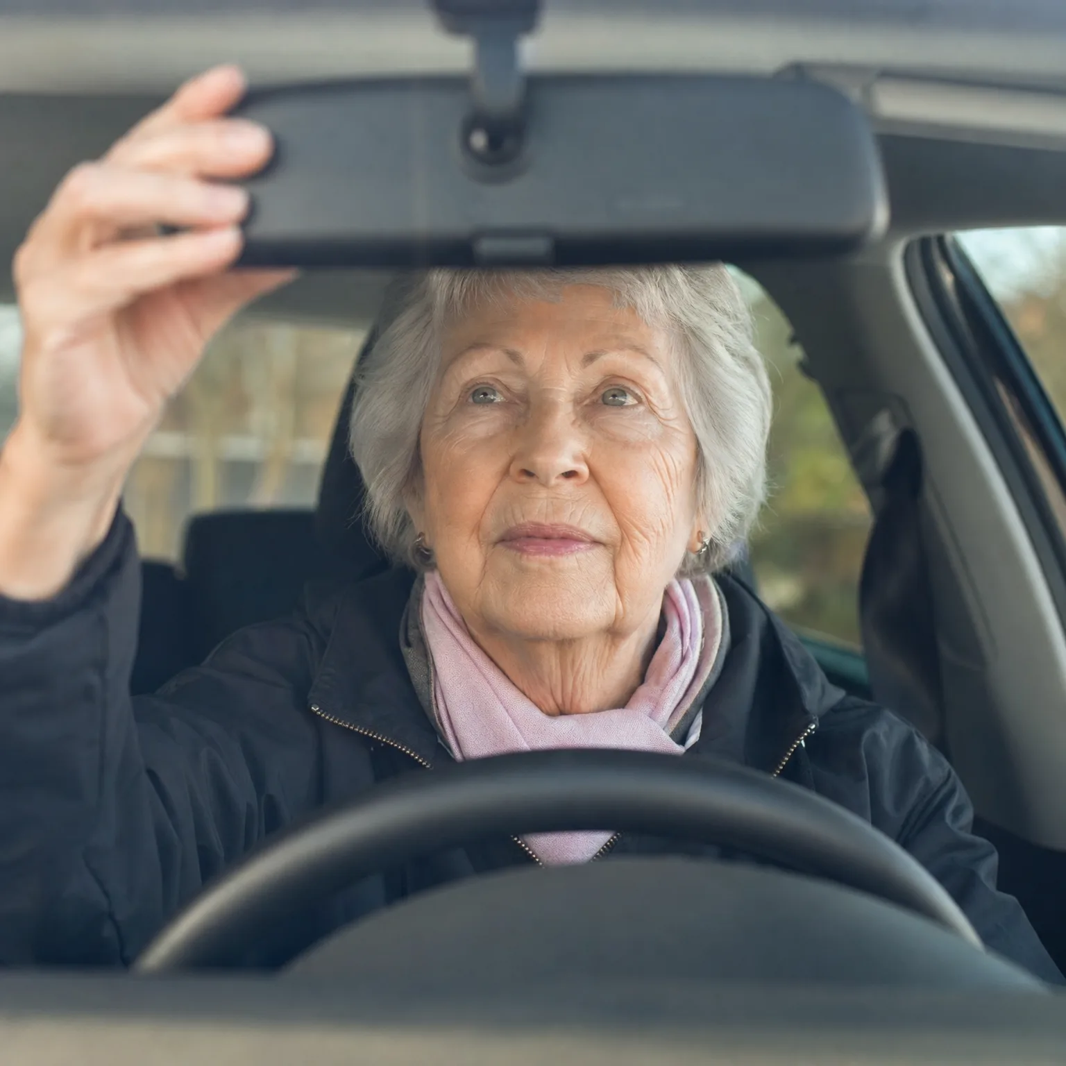 An older woman with short grey hair, wearing a dark jacket and pink scarf, sits behind a car steering wheel and reaches up to adjust the rear-view mirror.